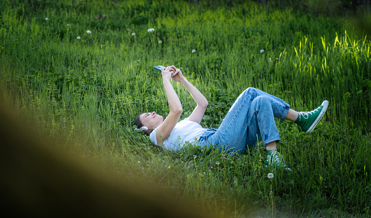 Young cheerful woman in a white t-shirt lying on the green grass in the garden looking into the smartphone screen.