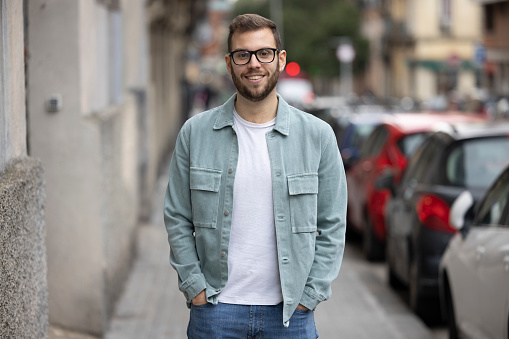 Young man, generation Z, standing in urban street scene in Barcelona, Spain, looking into camera, smiling friendly. He is wearing a turquoise shirt, a beard and black glasses. He might be a coworker or an entrepreneur in a start-up. In the background we see houses, traffic lights and parked cars.