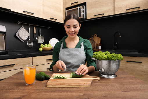 Woman in apron cutting cucumber at wooden table indoors