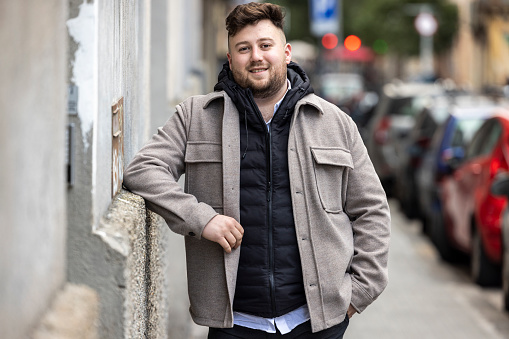 Young man, generation Z, standing in urban street scene in Barcelona, Spain, smiling friendly, cars parked in the background. He is wearing a beard, beige jacket and a hoodie, looking confident into camera.