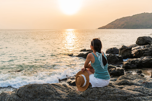 Rear view of beautiful young Asian woman sitting on stone enjoy moment of sunrise or sunset at the beach. Summer, vacation and holiday concept. Copy space