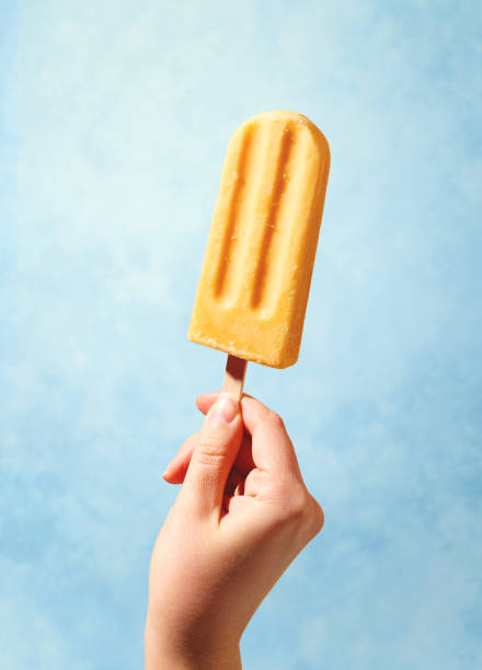 Woman hand holding yellow fruit popsicle on blue background Woman hand holding yellow fruit popsicle on blue background. Sunny summer mood popsicle stick stock pictures, royalty-free photos & images
