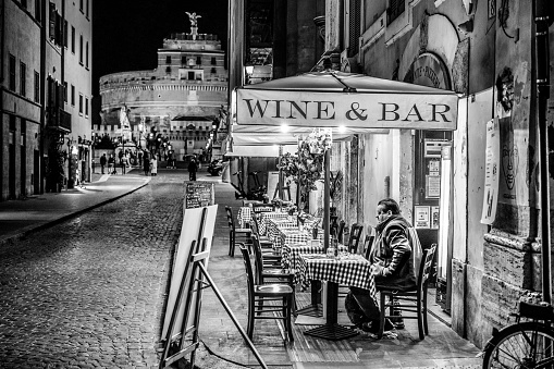 Rome, Italy, November 30 -- A lonely adult man sitting in the evening in a typical restaurant and wine bar along Via del Banco di Santo Spirito, in the Rione Ponte, in the historic heart of Rome. In the background the famous Castel Sant'Angelo. In 1980 the historic center of Rome was declared a World Heritage Site by Unesco. Image in high definition quality.