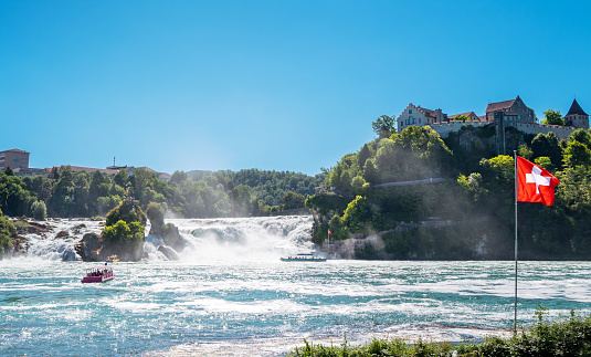 Schaffhausen, Switzerland - July 11, 2022: Early morning View of the Rhine Falls and Rhine River from the Schaffhausen promenade. Tour boats taking tourist to observation point.  The Schloss Laufen Castle in the background.