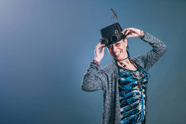Middle-aged woman with alternative style holds her hat while looking at camera with smiling expression in study shot on blue background