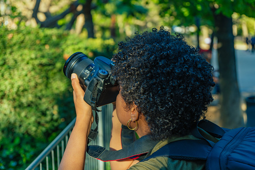 Young black African American woman with dark curly hair photojournalist taking a picture in a green park adjusting the focus ring and parameters of the photo camera