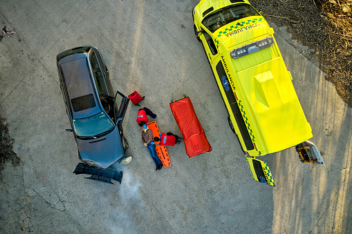 Drone point of view directly above car crash and emergency services team preparing man for transfer to ambulance and then hospital.