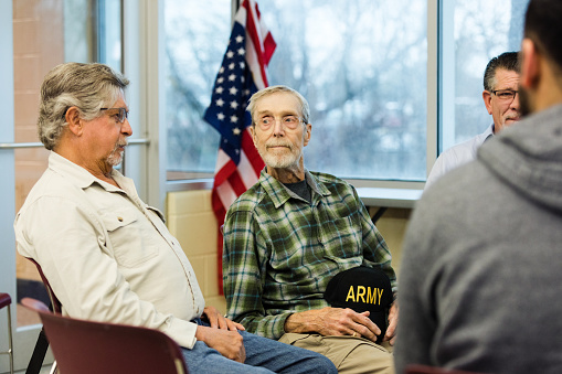 The senior adult man holding his army cap in his lap listens to a fellow Vietnam veteran at the meeting in the community center.