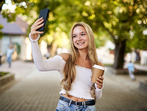 Young teeanager girl walking on a town street and taking selfie while holding takeaway paper coffee cup in her hand - Smiling student using her smartphone with headphones on her neck