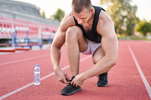 sportsman tying sneakers - man stopping lacing shoe outdoors - athletic shoes concept - a plastic bottle of mineral water in the background - playing field effort outdoors human age imagens e fotografias de stock