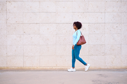Pretty woman with afro curly hair dressed in jeans, blue sweater and shoulder bag walking in the city with copy space on the left.