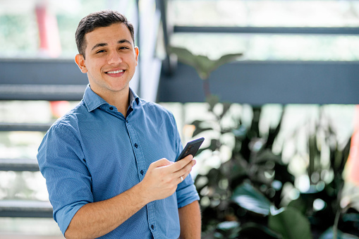 Portrait of a young businessman smiling while texting on his mobile phone by steps in the lobby of a modern office