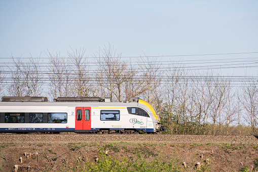 Side view of a Belgian electric train driving on the tracks with electrical wire above the train and trees in the background. public transport, transportation, railway embankment. 2 mei 2023, Eppegem