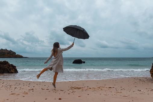 A young woman leaps in the air while grasping a black umbrella in her hand. Sesimbra, Portugal.