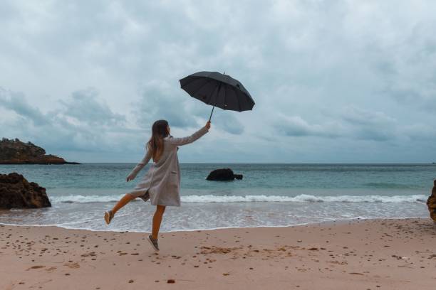 young woman leaps in the air while grasping a black umbrella in her hand. sesimbra, portugal. - white mid air rain wind imagens e fotografias de stock