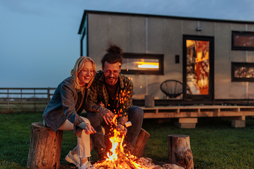 A romantic Caucasian couple is sitting at the campfire, roasting marshmallows in the evening on their vacation out of town.