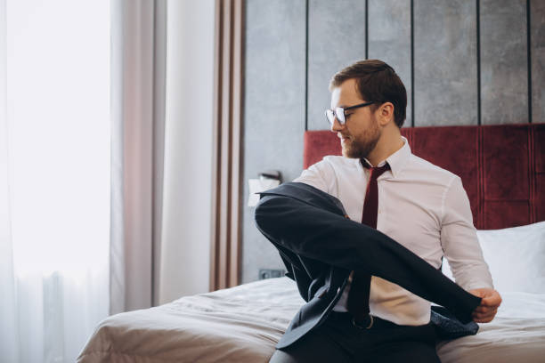 istock Business man with suitcase taking off jacket while sitting on bed in hotel room 1487351247