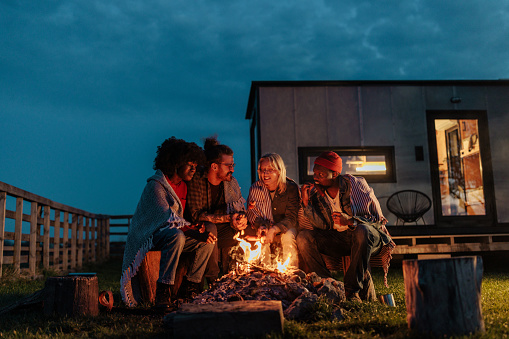 A group of young friends are camping in the nature at their cabin, roasting marshmallows by the campfire in the night.