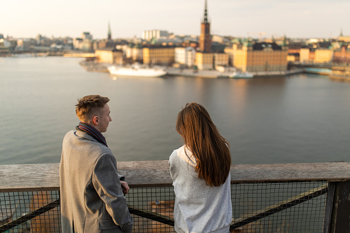 Teenage boy and girl standing on viewpoint above the Stockholm city. Wonderful view of the sea and cityscape at sunset. Two young people are talking and smiling.