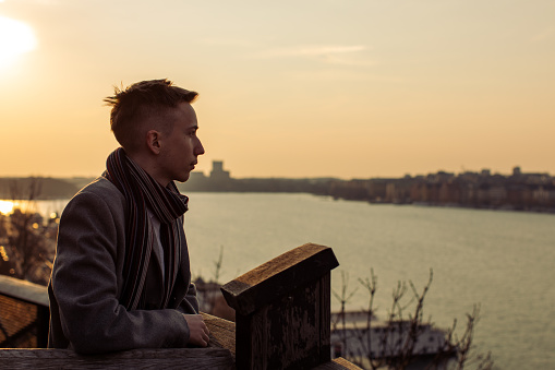 Teenager elegantly dressed in a coat and scarf, stands on viewpoint park balcony above the city. Boy leaning on fence enjoying a wonderful view of the sea and cityscape at sunset.