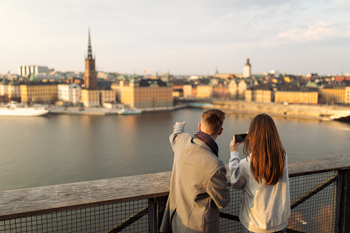 Teenage boy and girl standing on viewpoint above the Stockholm city. Boy is showing and pointing to the city while girl is taking photo with mobile phone. Wonderful view of the sea and cityscape at sunset. Two young people are talking and smiling.