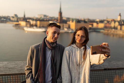 Teenage boy and girl standing on viewpoint above the Stockholm city. They are making a selfie with mobile phone with city in background. Wonderful view of the sea and cityscape at sunset. Two young people are talking and smiling.