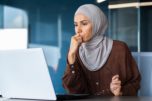 Portrait of a serious young woman in a hijab sitting in front of a laptop, holding glasses, thoughtfully and intently looking at the monitor.