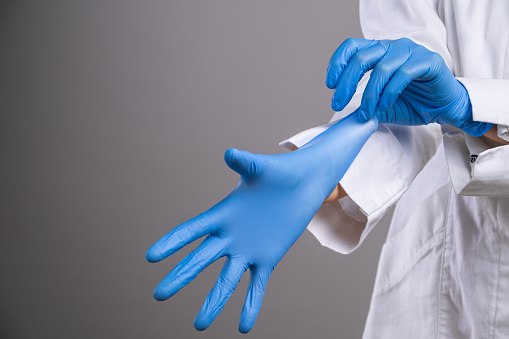 Close-up of a doctor, wearing a white coat, putting on a pair of blue surgical gloves.