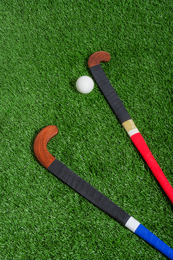 Looking down on a pair of wooden field hockey sticks wrapped one with red and the other with blue athletic tape with a white ball sitting on the turf.