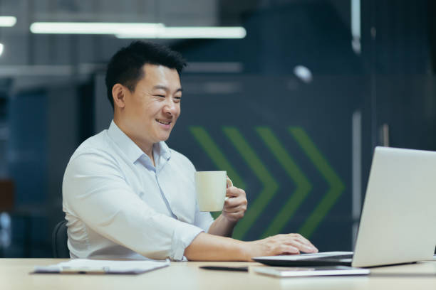 istock Asian smiling young man working in office on laptop, holding cup in hand, coffee break 1487359045