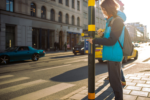 Teenage boy waiting at the intersection to cross the street. Boy in blue jacket presses the button on the traffic light to activate the green light for save crossing road, and eating his sandwich.