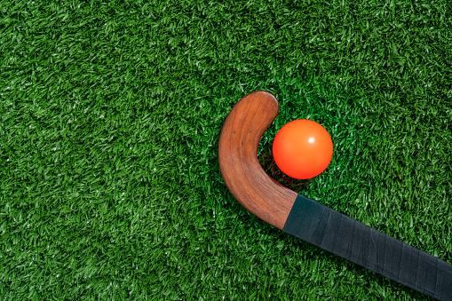 Looking down on a wooden field hockey stick wrapped with black athletic tape with an orange ball sitting on the turf.