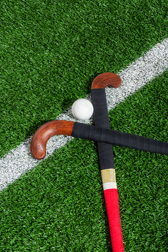 Looking down on a pair of wooden field hockey sticks wrapped one with red and the other with blue athletic tape with a white ball sitting on the white center line of a synthetic turf.