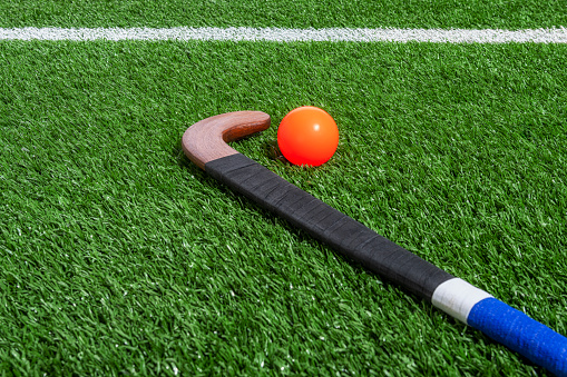 Low angle view of on a wooden field hockey stick wrapped with black and blue athletic tape with an orange ball sitting on the turf, with the centerline in the background.