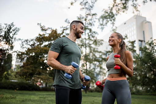 A happy sporty couple doing exercises for biceps with weights outdoors
