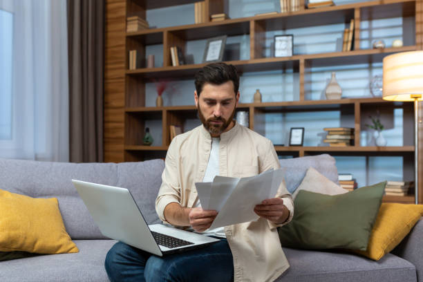 istock A young man works remotely with documents in quarantine. Sitting on the couch, holding a laptop and looking at papers, bills 1487362554
