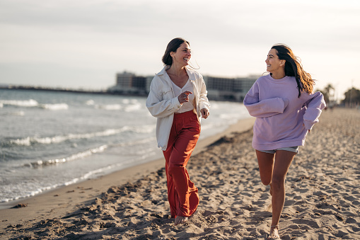 Two beautiful lovely young women feeling happy and carefree running with joy barefoot on sandy beach by the ocean, having great time, looking at each other, smiling, enjoying early morning routine.