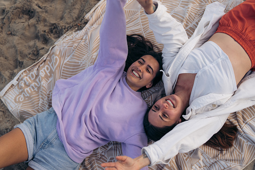 View from above of two young female friends tourists lying on picnic blanket on sandy beach with arms outstretched, smiling, having fun, enjoying vacation, chilling outdoors, looking happy and relaxed.