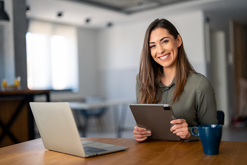 Smiling confident businesswoman looking at camera sitting at home office desk. Modern stylish female manager, successful female entrepreneur holding digital tablet posing for business portrait.