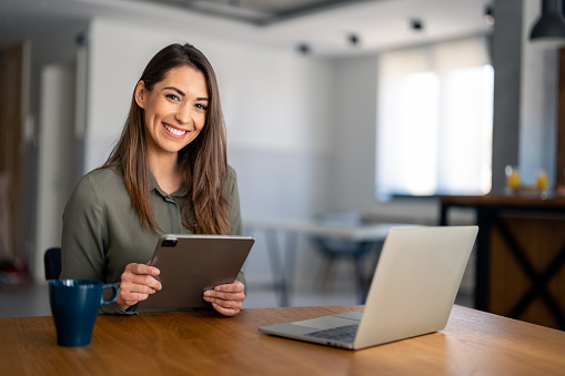 Portrait of smiling fulfilled young woman holding digital tablet device, looking at camera, sitting at desk at home with laptop computer in front of her and cup of coffee.