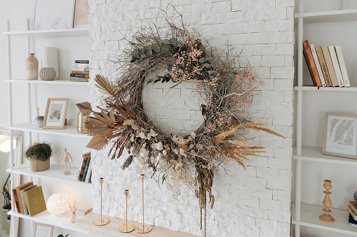 A wreath of dried flowers over the fireplace between shelving with books and decor in a stylish modern Scandinavian living room. Nobody.