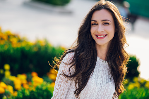 A cheerful chic boho girl is standing in city park and smiling at the camera. Beauty photo of a young happy brunette with thick and lush hair making eye contact.