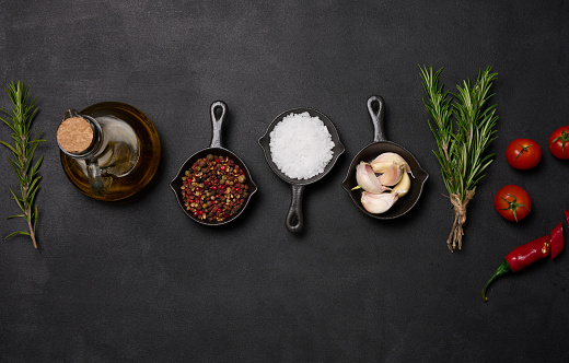 Miniature pans with spices, salt, black pepper and fragrant pepper, a sprig of rosemary on a black table. Spices for cooking