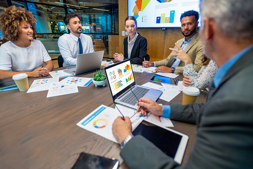 Diverse group of people meeting and working at a board room table at a business presentation or seminar. The documents on the conference table have financial or marketing figures, graphs and charts on them.