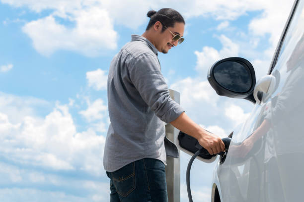 Progressive man with plugged-in EV car with charging point, cloudscape backdrop. Progressive man with electric vehicle and cloudscape background. EV car driven by clean renewable plugged-in with charging point. Refreshing environment from green energy. happy car asian stock pictures, royalty-free photos & images