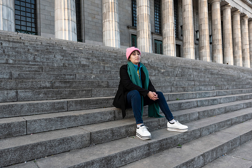 Female latin student with brown hair and pink hat sitting on the stairs in college building