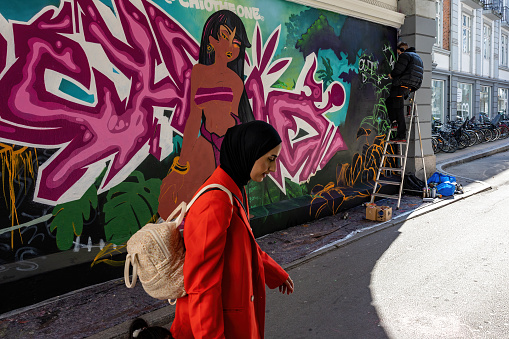 Copenhagen, Denmark April 9, 2023 A Muslim woman dressed in red passes a mural in a passageway with the artist..