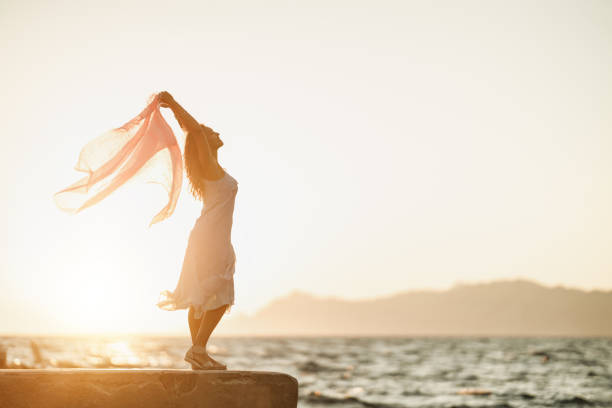 An attractive young woman is having fun and enjoying a summer vacation. She is posing with transparent scarf in her hands on the beach.