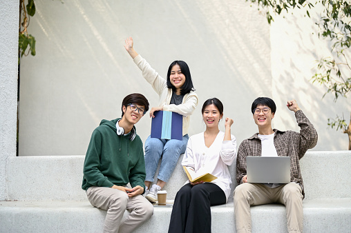 Image of happy and cheerful Asian college students sitting on a bench at a campus relaxation area together. Friendship, togetherness, university lifestyle