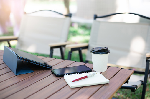 Cup of coffe and smartphone, tablet and note book on wooden table with lawn chair. coffee break and relax after work in parks. No people, Blurred  background